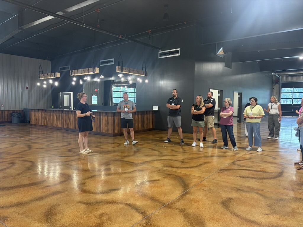 A group of people stand in a room with a wooden counter, facing a woman speaking. The room has patterned flooring and metal beams on the ceiling.