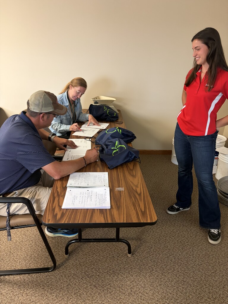 Three people in a room, one seated with paperwork, another standing, and a third seated, all engaged in discussion.