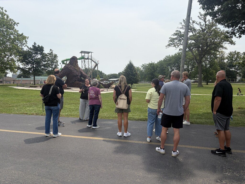 A group of people in casual clothing stand in a parking lot. Behind them is a grassy area and trees.