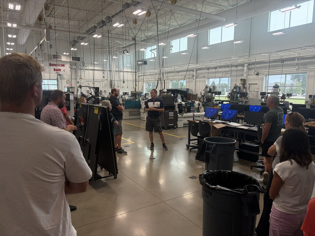 A group of people in a factory setting with machines, a trash can, and overhead lighting.