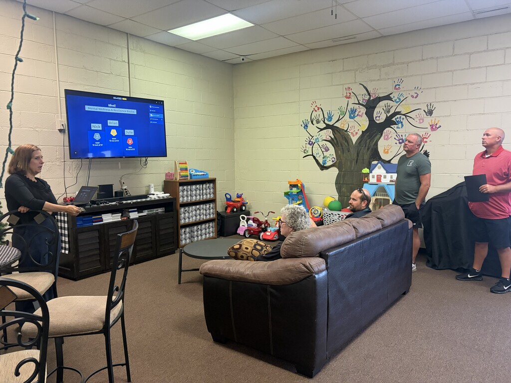 Four people in a room with a black leather couch, large screen, chairs, a tree mural, and a woman standing by a computer.