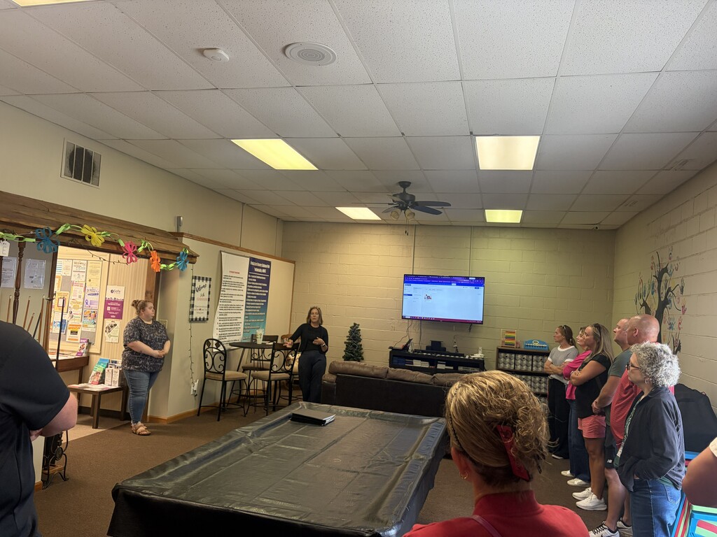 A group of people stand around a room with a woman speaking. A large screen is mounted on the wall.