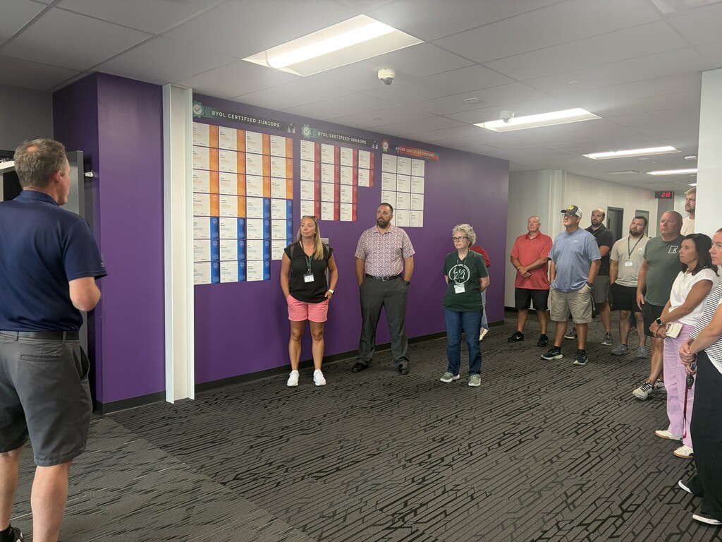 Several people in various clothing stand in a room with purple walls and a large bulletin board.