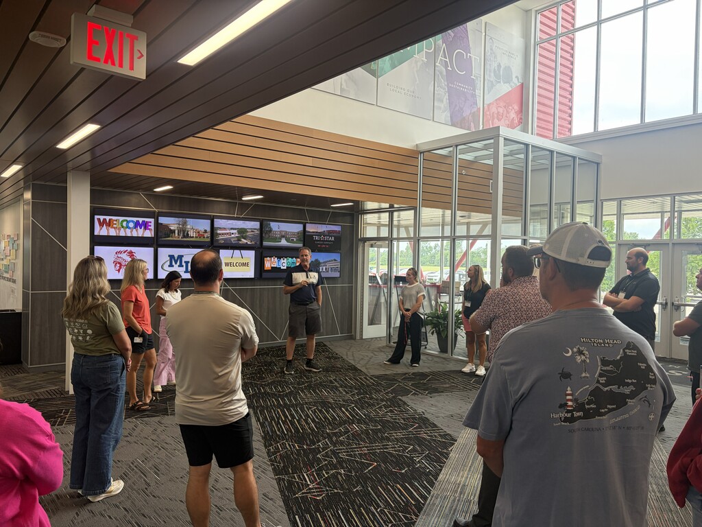 A group of people in a lobby with screens, some standing, and an "EXIT" sign overhead.