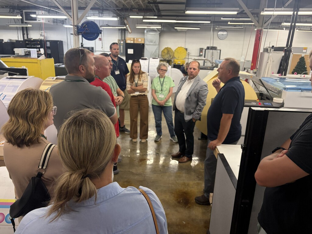 A group of people stand in a factory area, some in conversation, with machinery and equipment visible in the background.