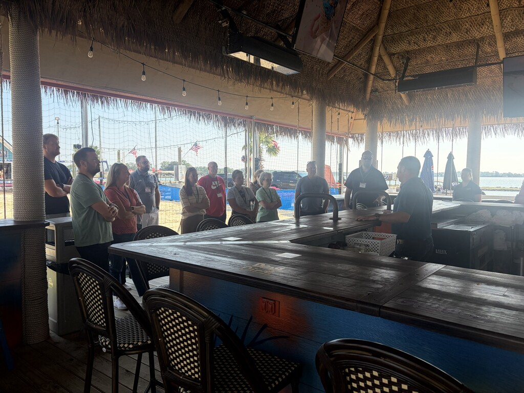 A bar with wooden counter and chairs. People stand and talk near the bar. An ocean is visible in the distance.