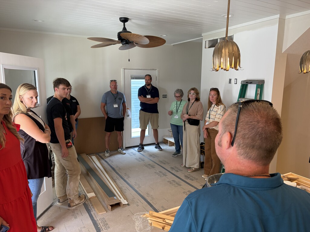 Group of people in a room with unfinished walls, a ceiling fan, and an open door.