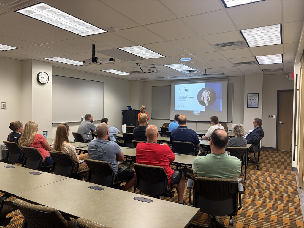 A classroom with participants seated around tables, a person at a podium, and a projector screen.