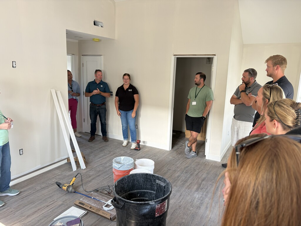 A group of people are standing in a room with bare walls, listening to a man in a green shirt.