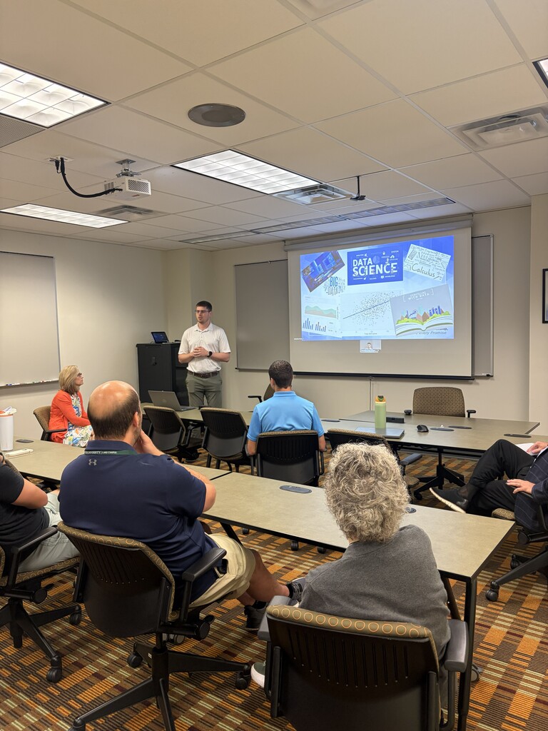 People seated at tables in a classroom, facing a man standing by a projector screen.