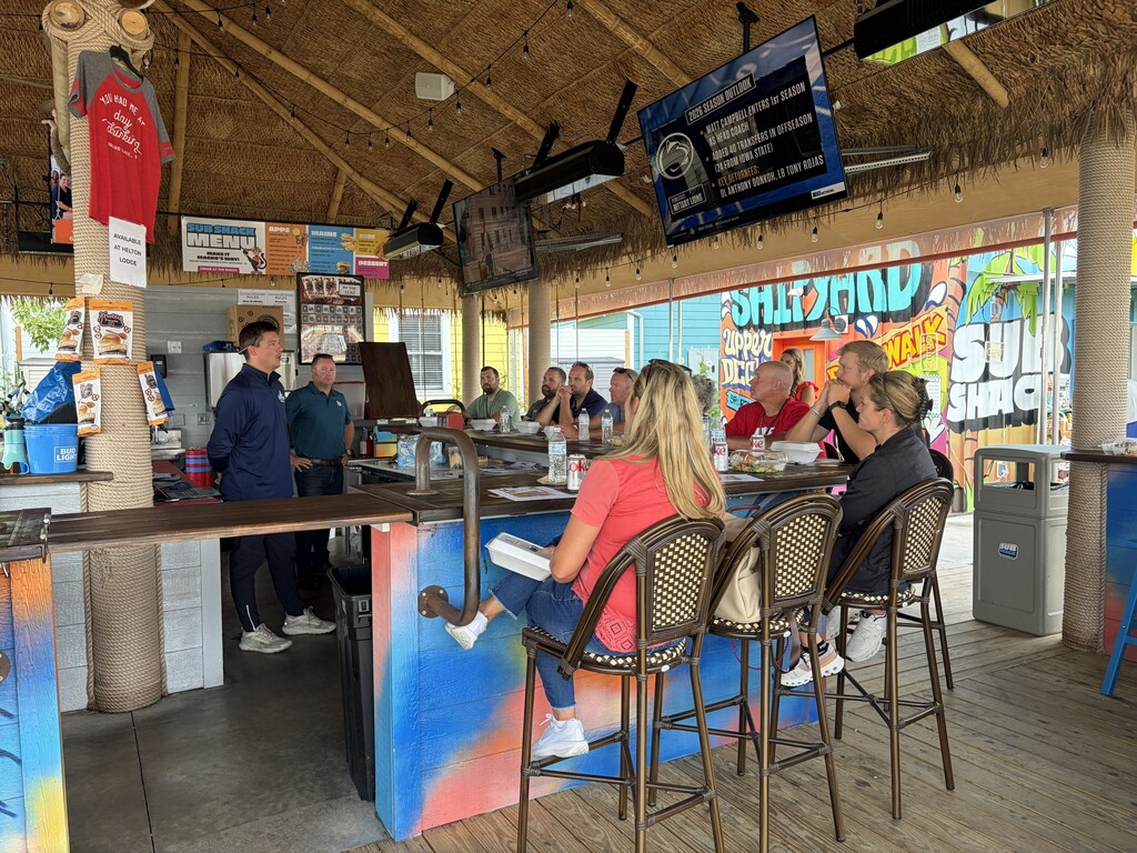 A group of people sit around a bar inside a thatched-roof restaurant. A bartender stands behind the counter.