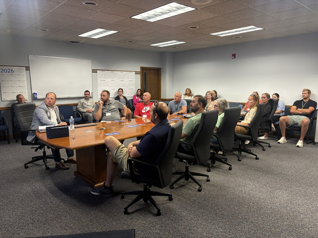 A conference room with a large table has people seated. Whiteboards and ceiling lights are visible.