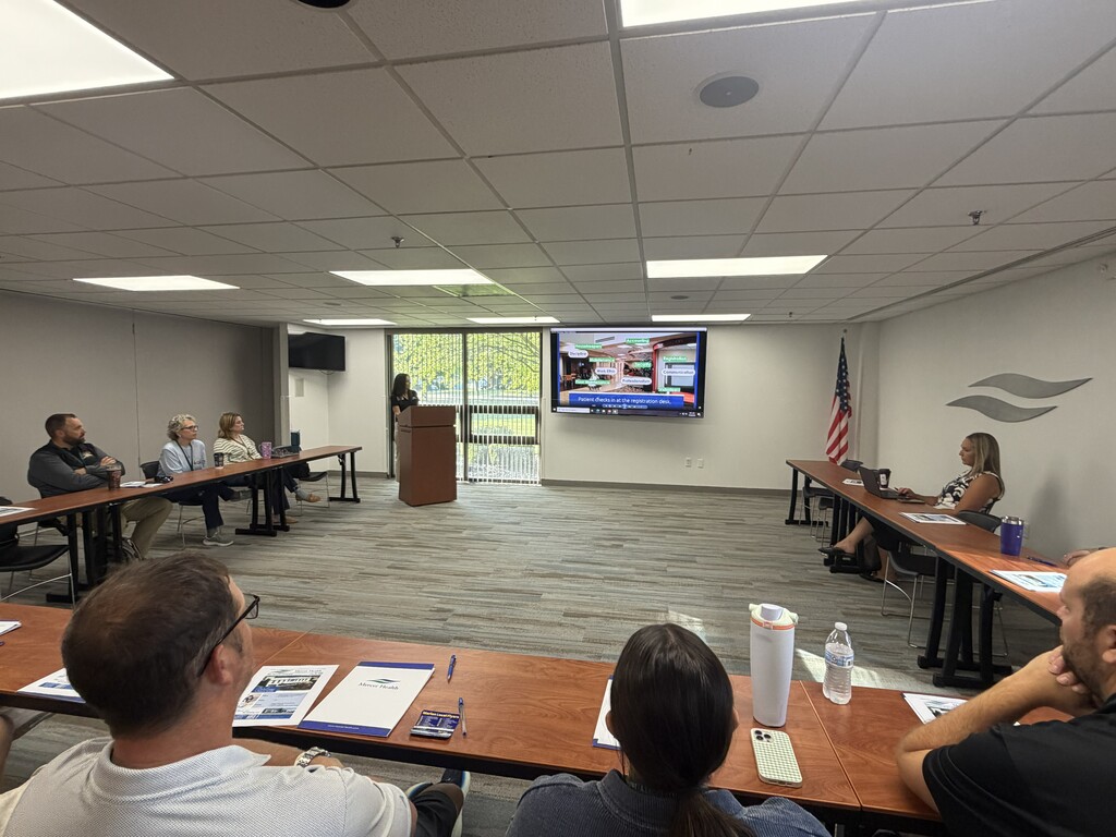 Meeting room with people seated at tables. A woman speaks at a podium. A TV screen is mounted on the wall.