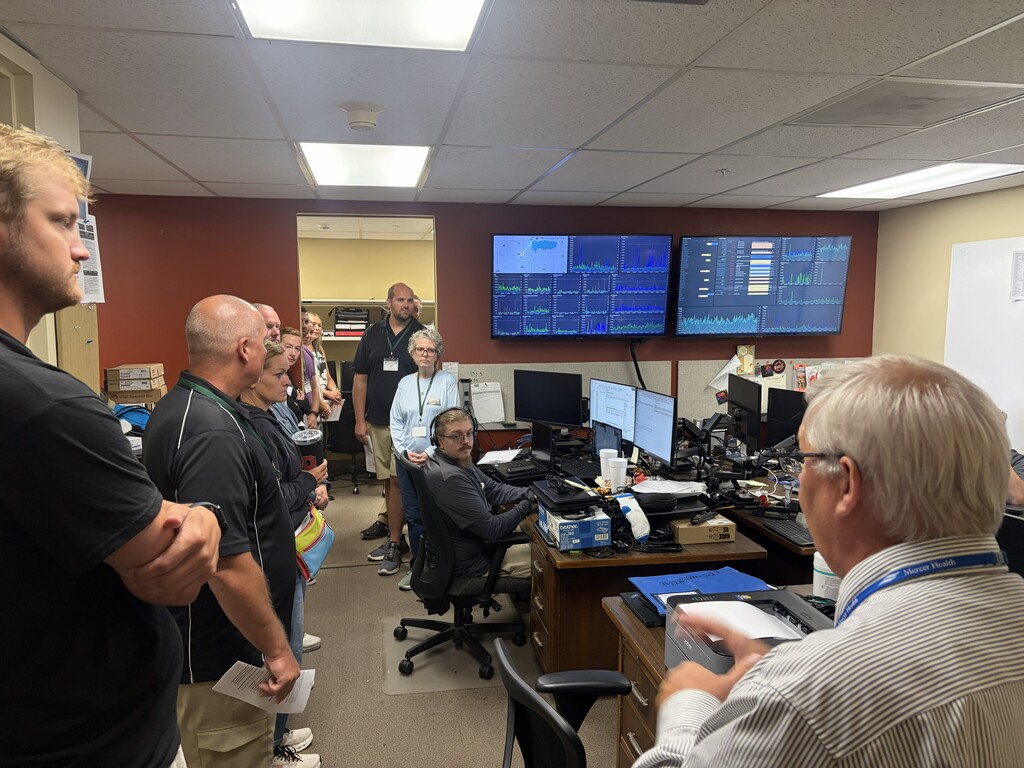 Several people gathered in an office. One man sits at a desk, facing a computer. Another man stands and speaks.