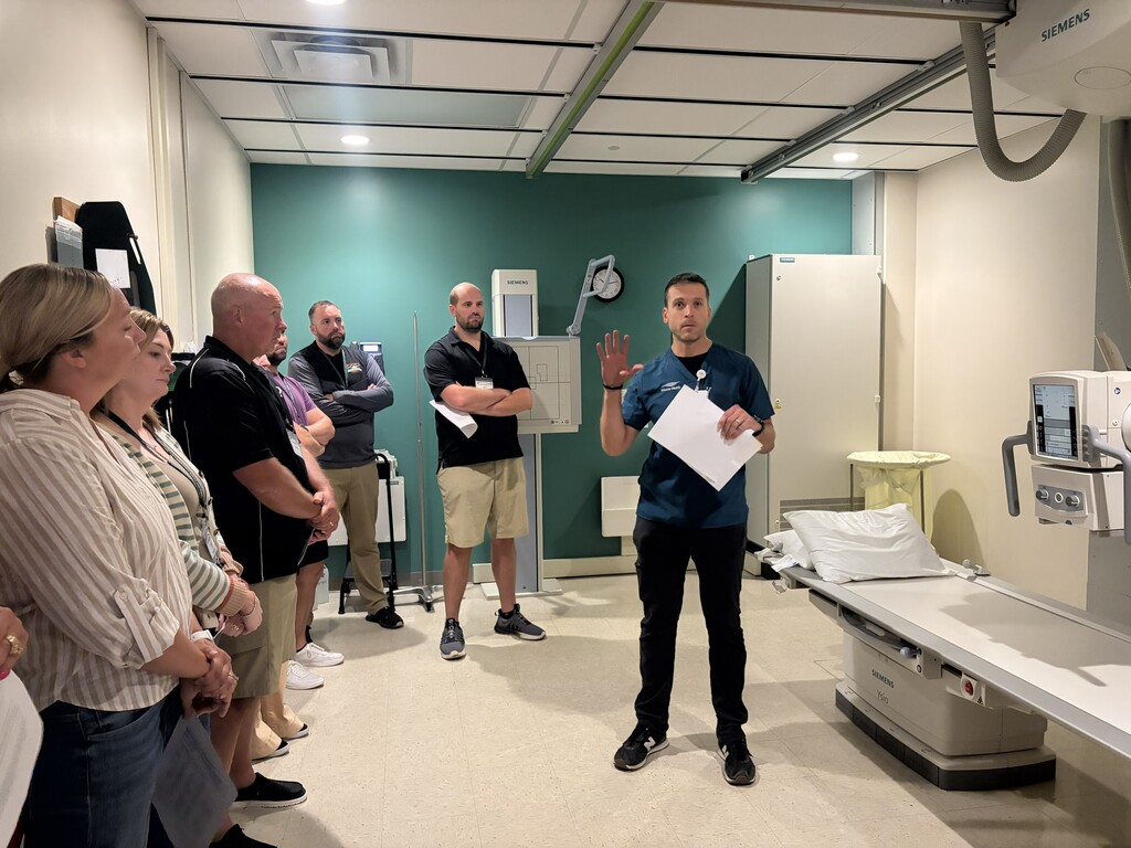 A man in a blue shirt stands in a medical room, speaking to a group of people.