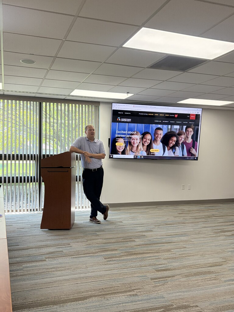 A person stands at a podium in a room with a large screen on the wall.