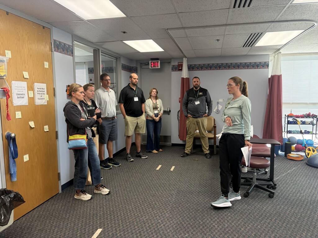 Several people stand in a room with carpeted floors, listening to a woman in white. Behind her, gym equipment is visible.