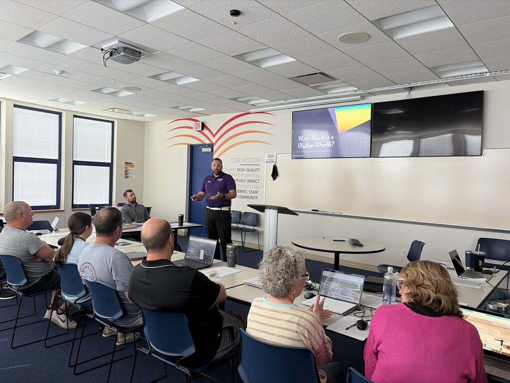 A classroom with several people seated in chairs, facing a person standing at a podium.