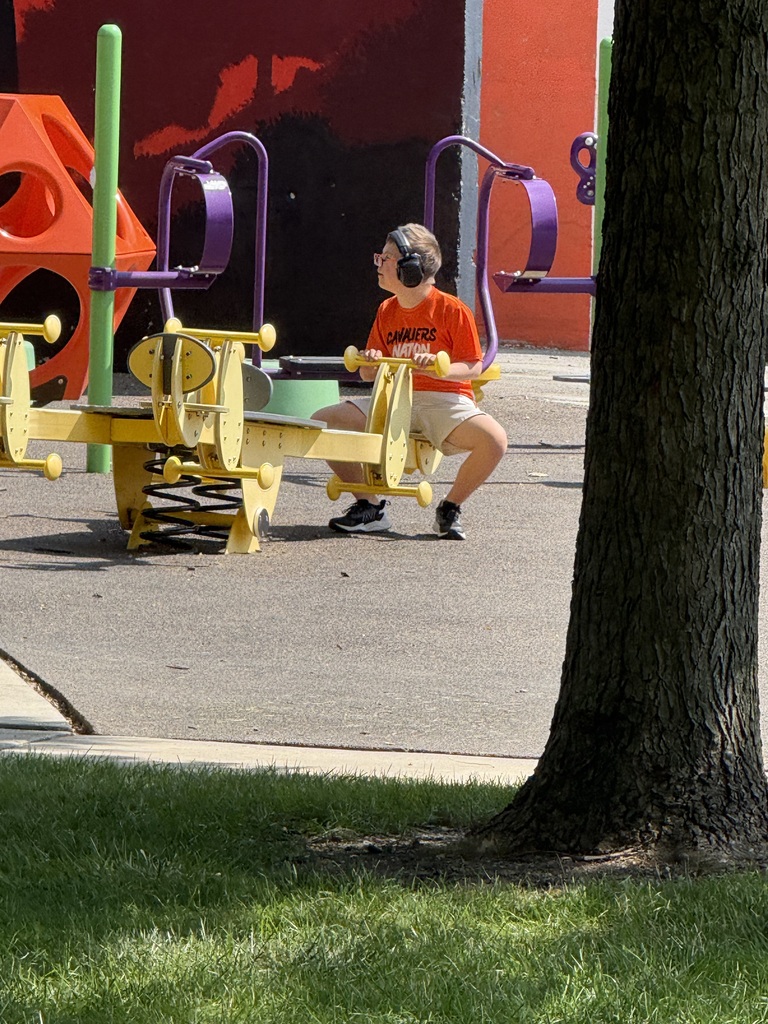A child wearing headphones and an orange shirt plays on a yellow playground structure with a red wall in the background.