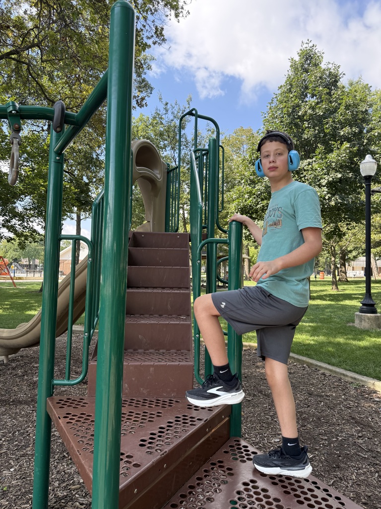 A child stands on steps at a playground, wearing headphones. Green railings and a slide in background.