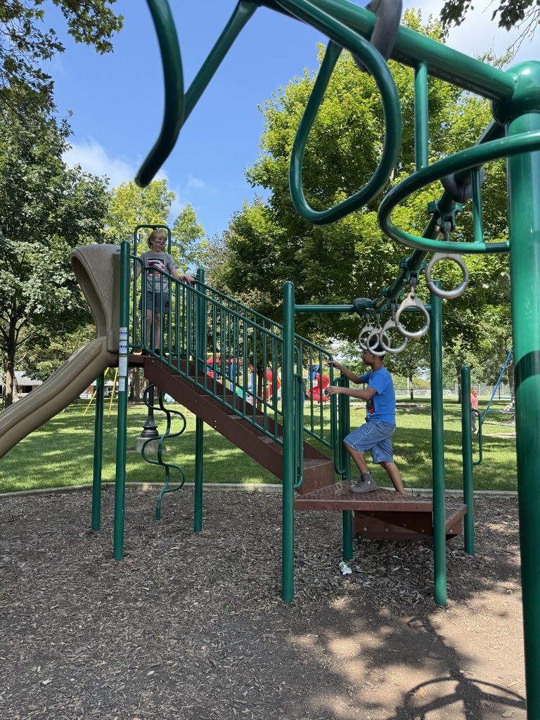 Two children are climbing a green metal structure with a slide and handrails in a park.