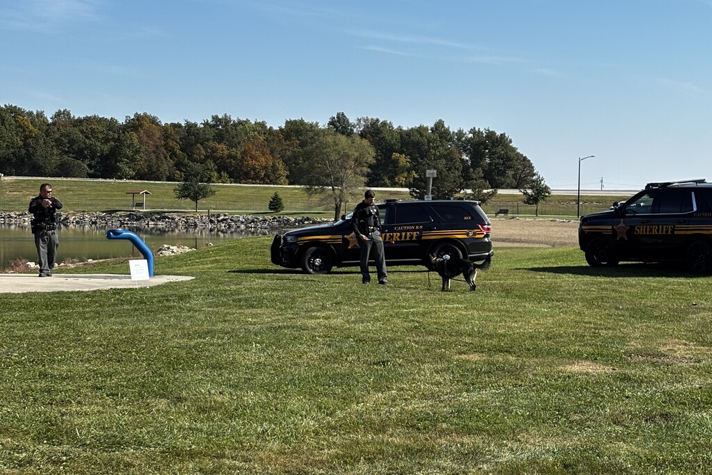 Three police officers with a dog on a grassy field. Vehicles parked. Trees and a building in the background.