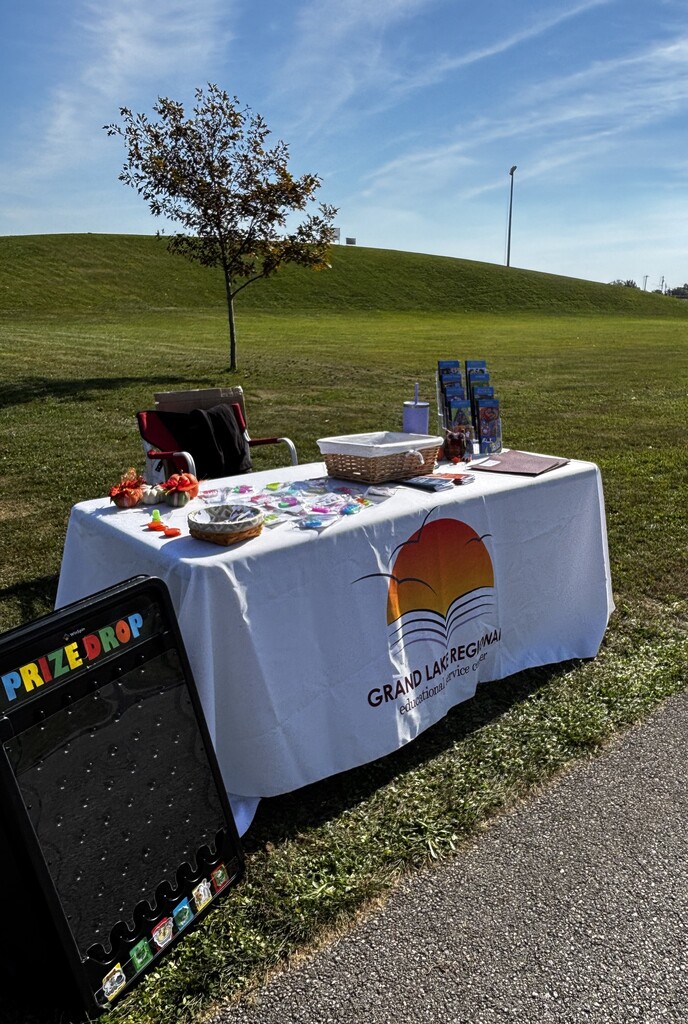 A table with a white tablecloth has a sign reading "Prize Drop" and several items on it. A small tree is behind the table.