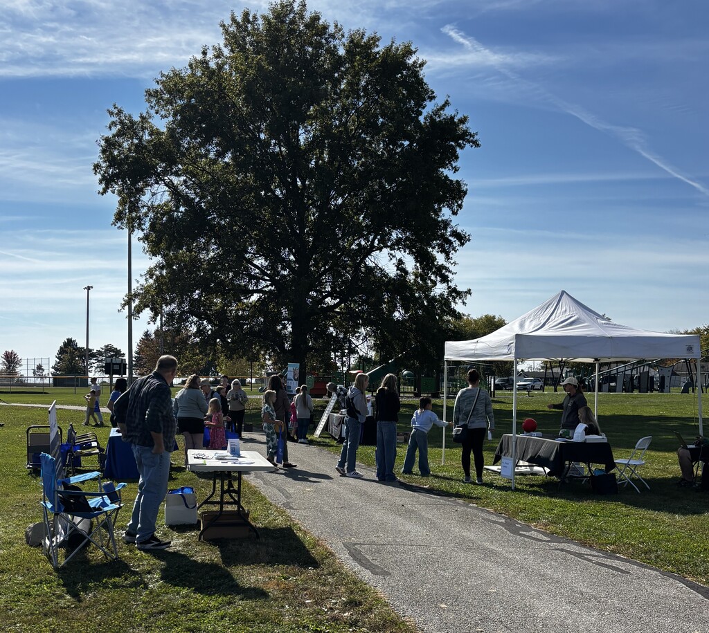 Group of people gather around tables and tents on a grassy field with a large tree.