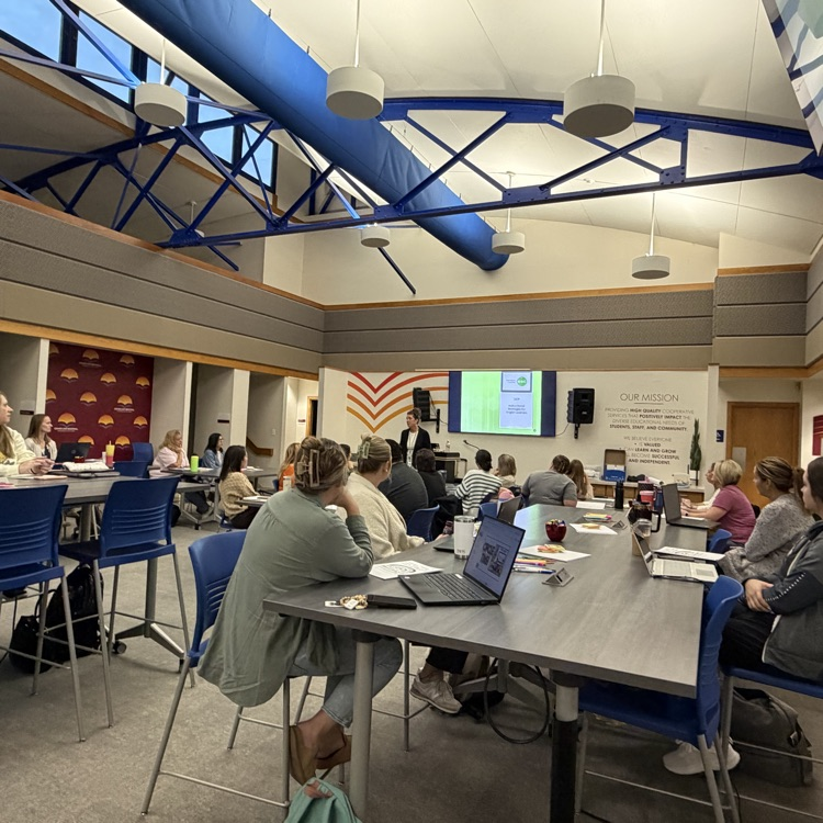 Students and teachers sit at tables with laptops and papers, a person standing at the front of the room near a projector.