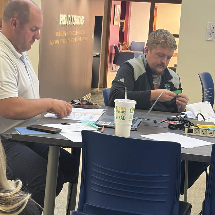 Two men seated at a table with papers, a cup with a straw, a cell phone, and a laptop.