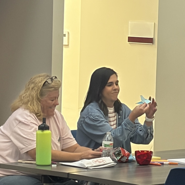 Two women seated at a table with papers, a water bottle, and a small red bowl.