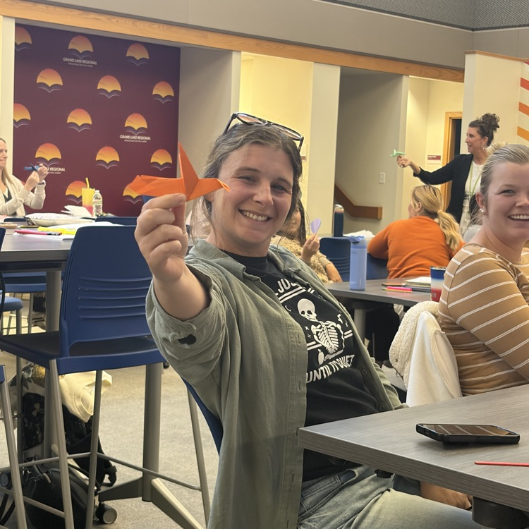 People sit around tables in a room. A woman holds an orange paper craft, smiling. Behind, a woman stands by a wall with designs.