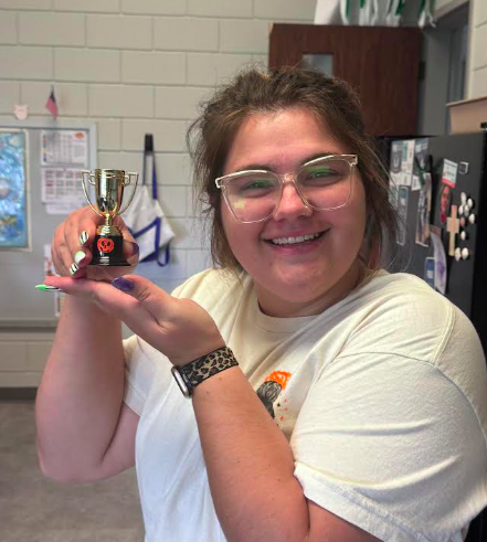 Person with glasses holds small trophy up in front of face. A refrigerator and bulletin board in background.