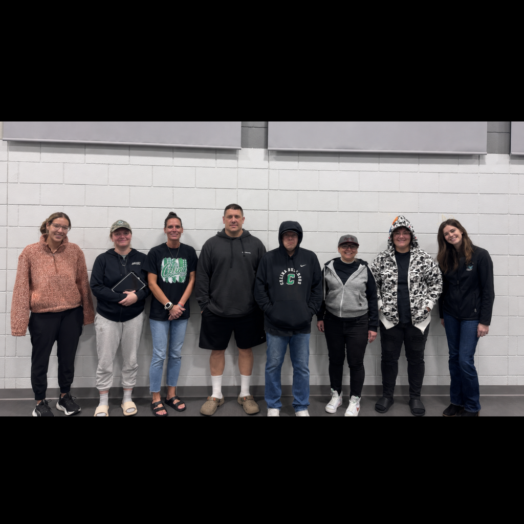 Seven adults standing in front of a white wall, each wearing different clothing and shoes.
