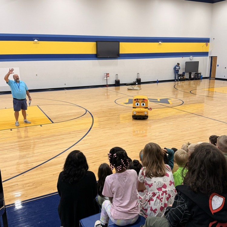 School gym with kids watching a yellow electronic bus give bus safety lessons  