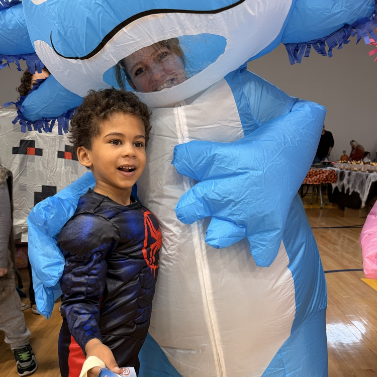 Little boy with dark hair in a super hero costume hugging an inflatable blue dinosaur  