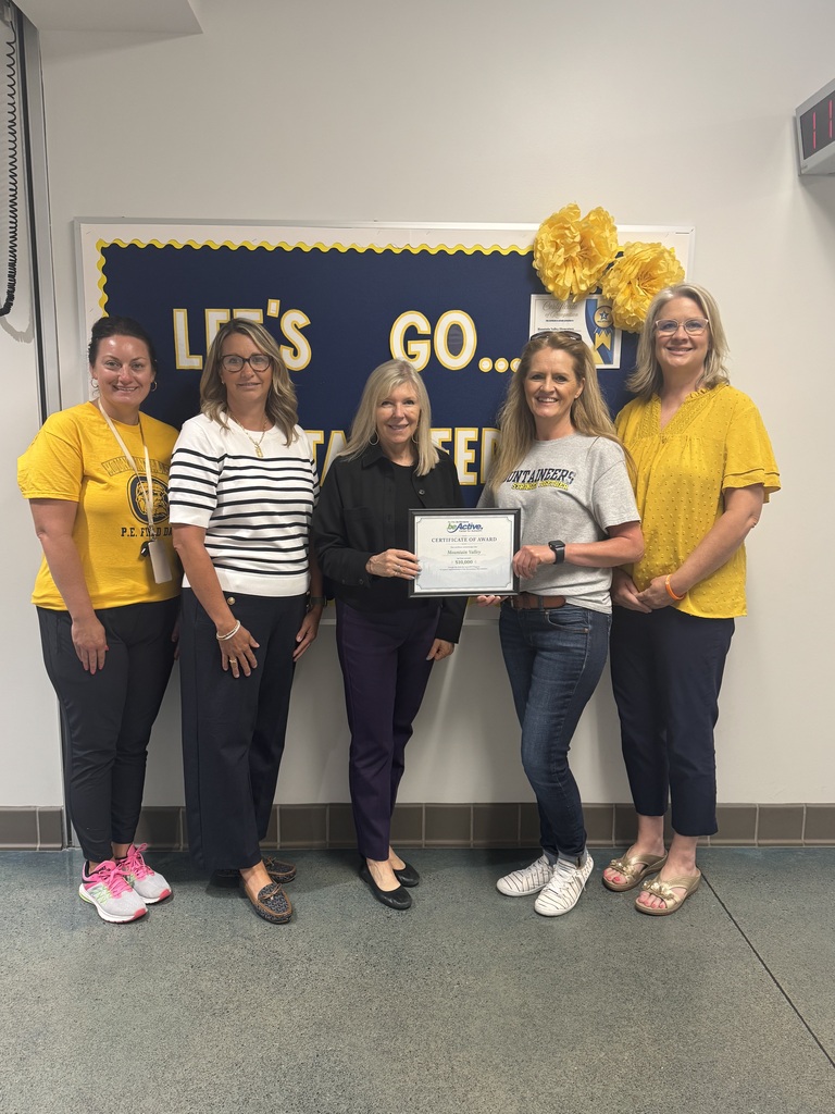 Five women in yellow shirts stand in a room with a blue banner and yellow flowers. The woman in the center holds a certificate.