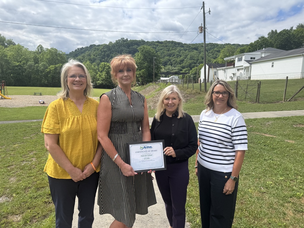 Four women stand on a path in a grassy area, holding an award. A house is visible in the background.