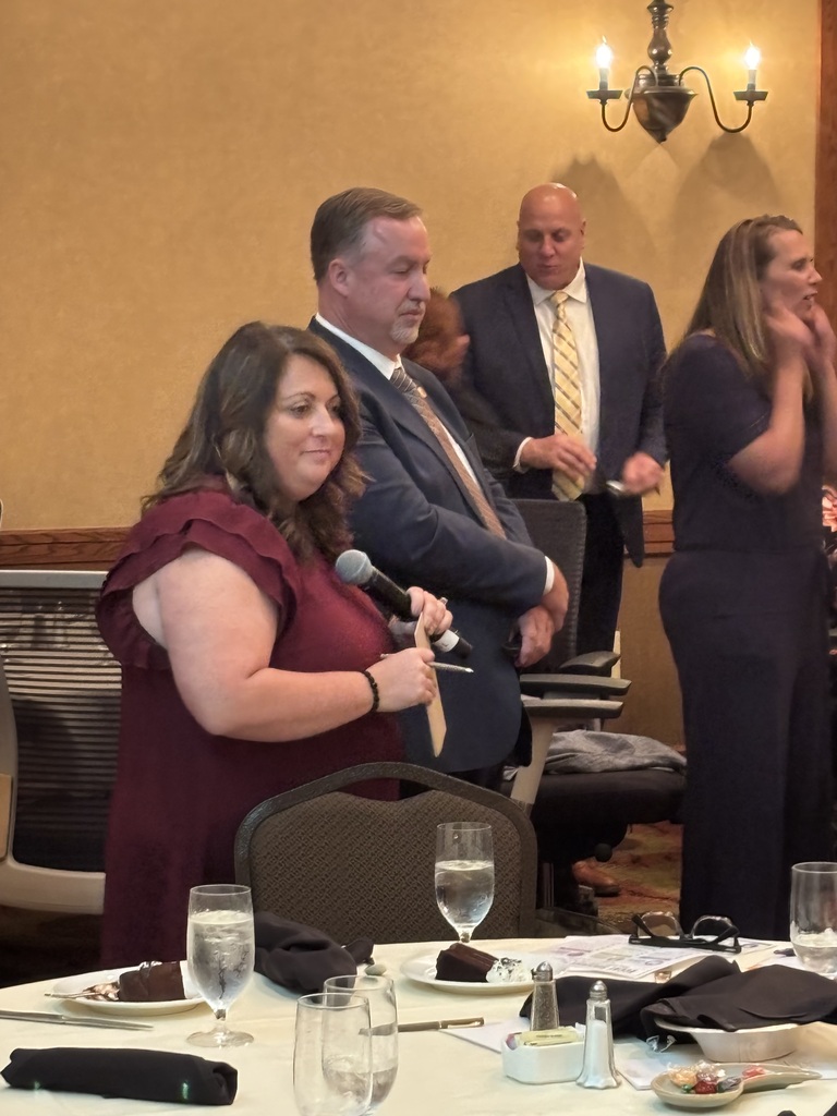 A woman speaks into a microphone at an event, with several people standing behind her. A table holds water glasses, a plate, and a napkin.