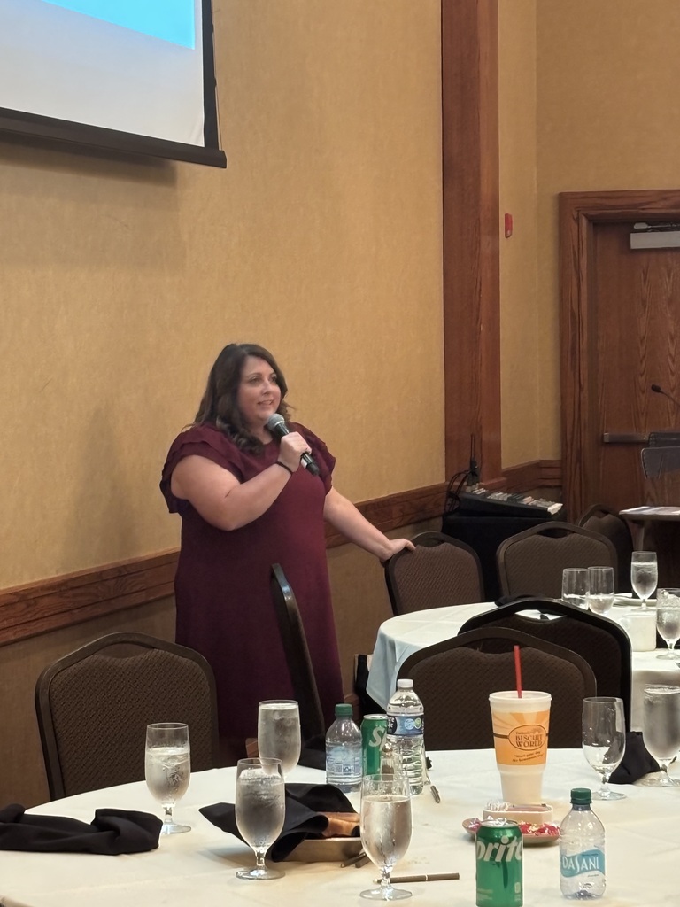 A woman speaks into a microphone in a conference room. There are tables and chairs in the background.