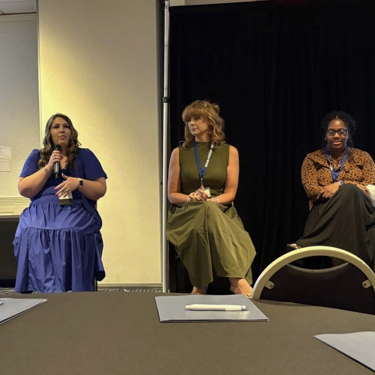 three women sitting on a stage. one woman is speaking into a microphone  
