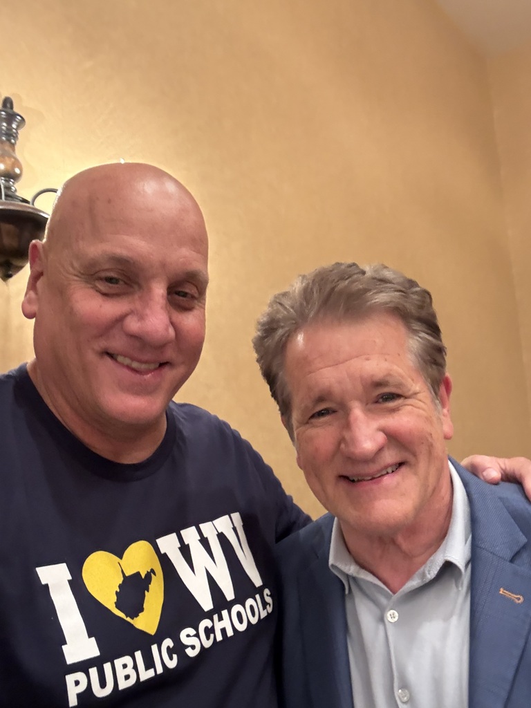 Two men stand close, smiling. One wears a dark blue shirt with "I [heart] WV PUBLIC SCHOOLS". The other wears a blue blazer. Background is light yellow.