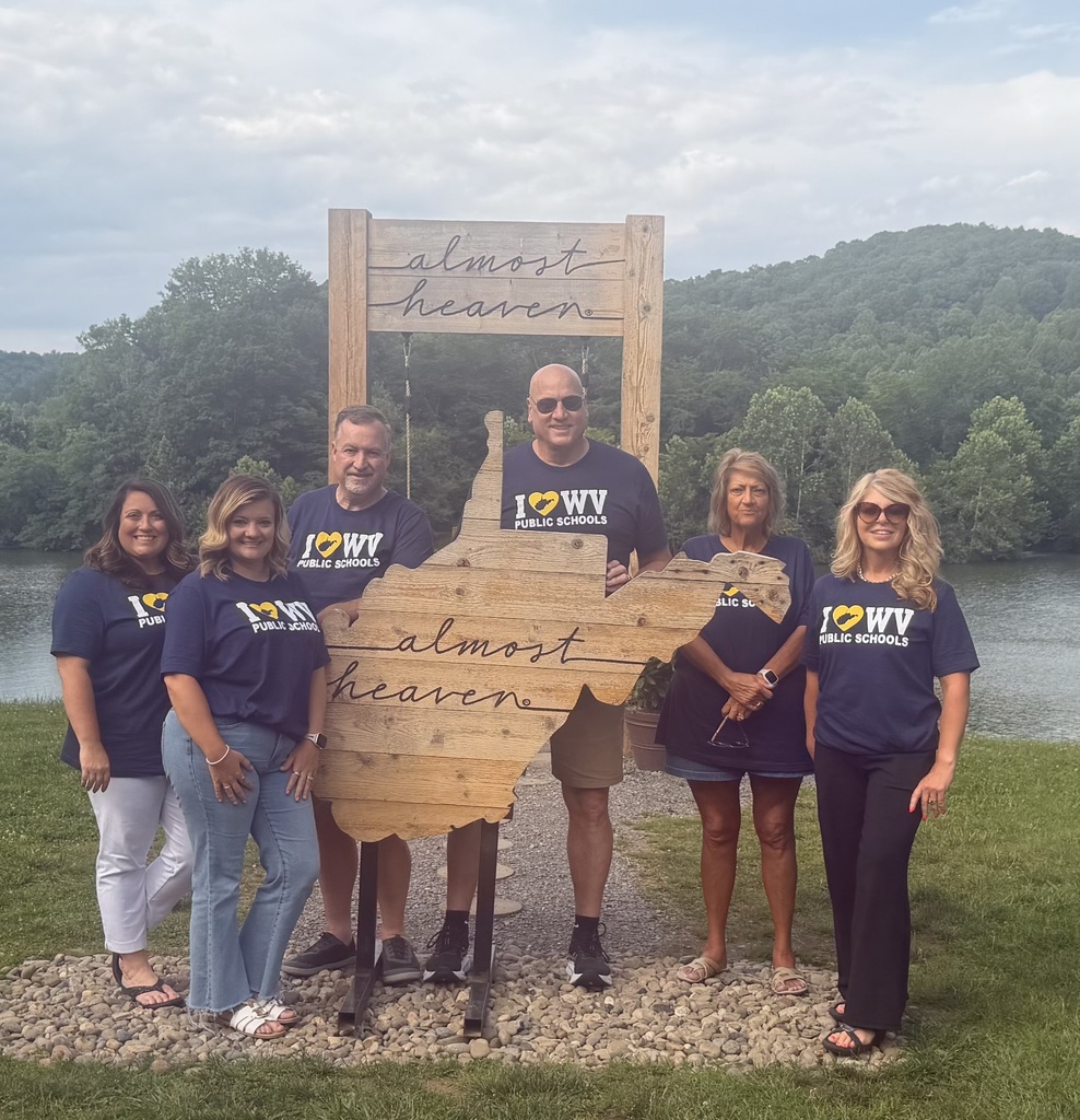 A group of six people in matching blue shirts stands in front of a sign with "Almost Heaven" and a West Virginia map.