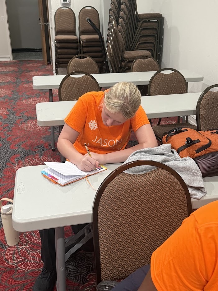 person in orange shirt seated at a table writing in a notebook