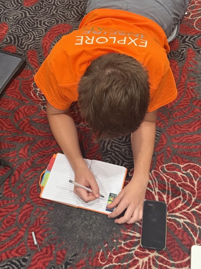 A person in an orange shirt lying on a carpeted floor, writing in a notebook with a pen.