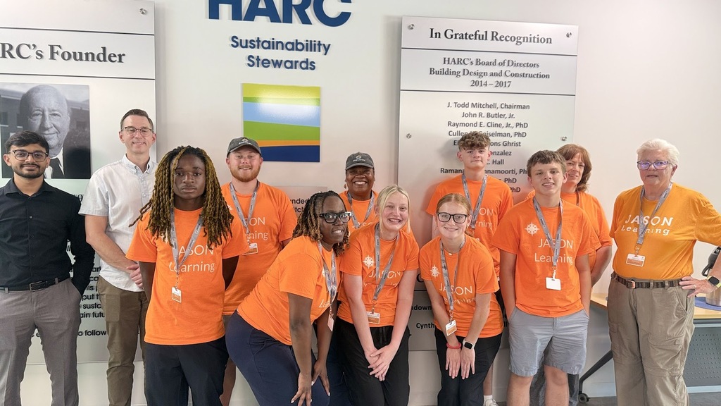 A group of people, some in orange shirts, stand together in front of a wall with a sign.