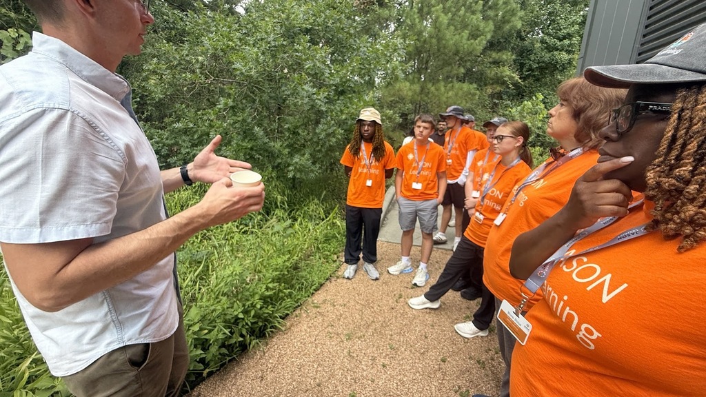 A group of people in orange shirts, listening to a man in a white shirt standing in a park.