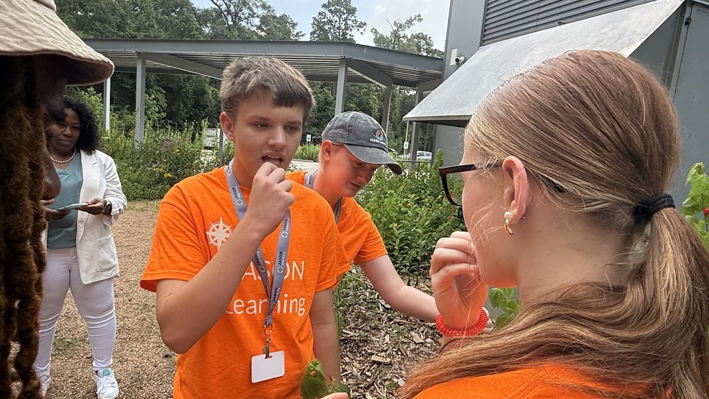 Four people in orange shirts, one of them with a microphone, are standing outdoors near plants and buildings.