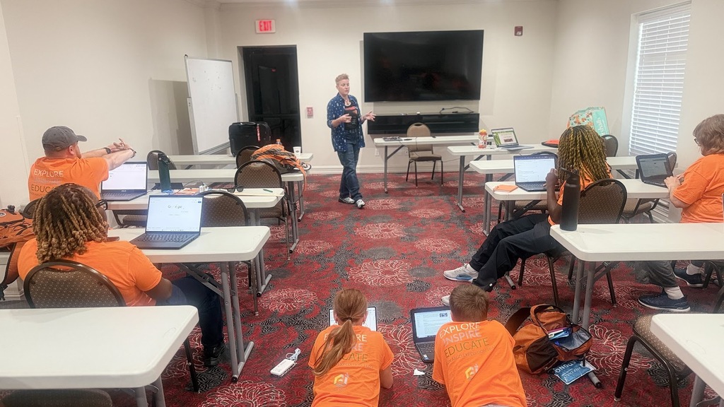 A classroom with students in orange shirts, seated at desks with laptops, and a person standing in front.