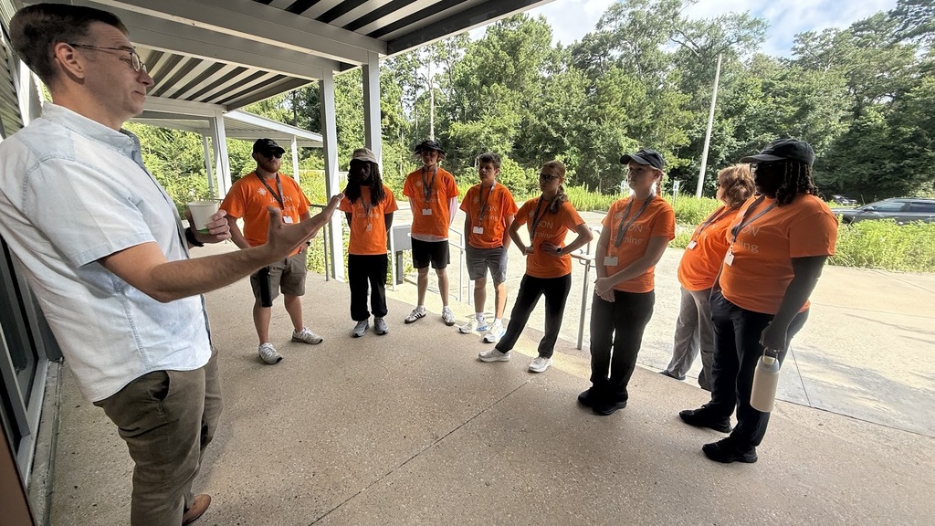A group of people in orange shirts gather, listening to a man in a white shirt speak.